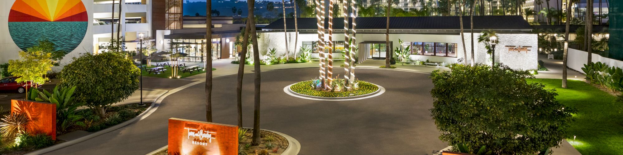 The image shows a hotel entrance with palm trees, landscaped greenery, and modern architecture, featuring a mural of a sunset on a building. Town and Country Resort is a hotel that has trolley access to Snapdragon Stadium, Viejas Arena, Petco Park, and the Rady Shell.