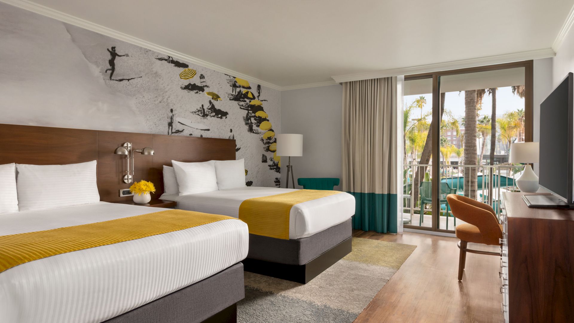 The image shows a hotel room with two beds, a wall mural, a TV, a chair, a lamp, and a balcony with a view of palm trees and buildings close to Snapdragon Stadium, Viejas Arena, and Petco Park so you can enjoy watching the San Diego State Aztecs football team, San Diego FC of Major League Soccer, and San Diego Wave FC of the National Women's Soccer League.