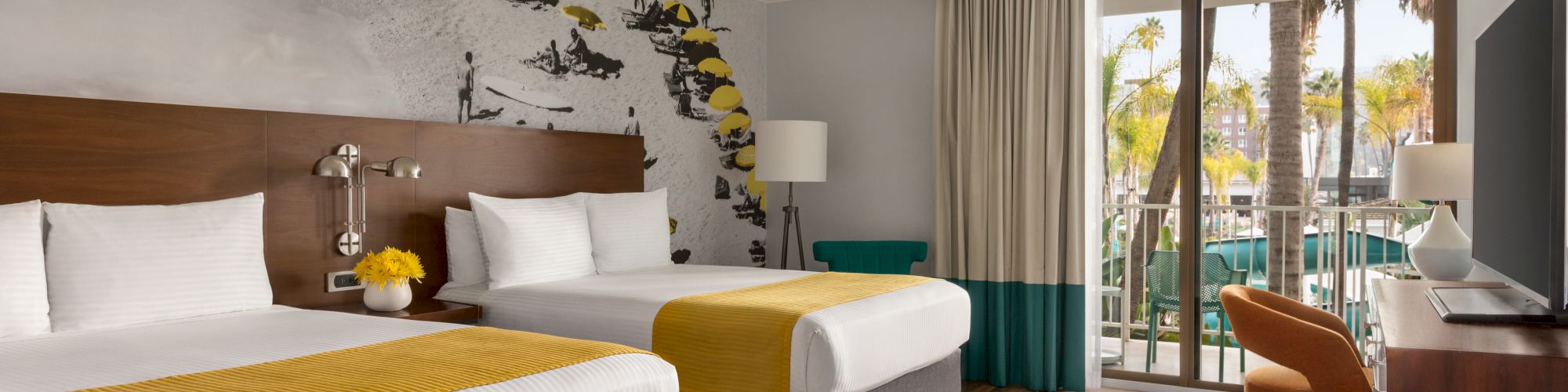 The image shows a hotel room with two beds, a wall mural, a TV, a chair, a lamp, and a balcony with a view of palm trees and buildings close to Snapdragon Stadium, Viejas Arena, and Petco Park so you can enjoy watching the San Diego State Aztecs football team, San Diego FC of Major League Soccer, and San Diego Wave FC of the National Women's Soccer League.