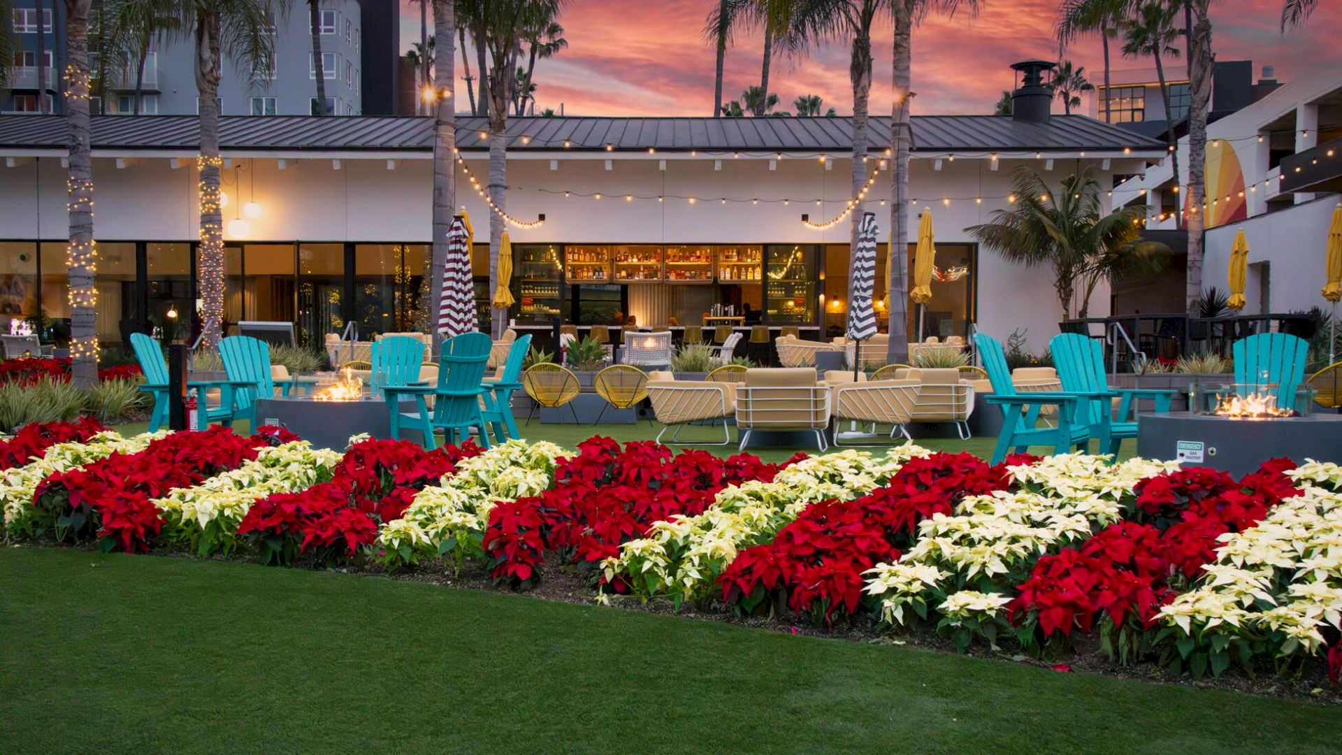 A vibrant outdoor lounge with colorful chairs, floral arrangements, and palm trees against a sunset sky.