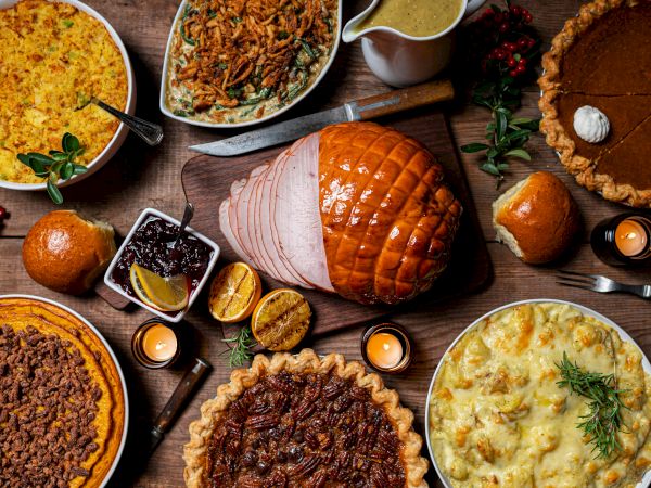 Rustic holiday feast with glazed ham, casseroles, gravy, stuffing, pot pie, bread, lemons, and cookies on a wooden table.