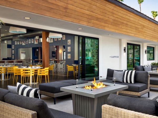 A modern patio lounge with cushioned seating and a lit outdoor firepit, beside a restaurant dining area and palm trees.