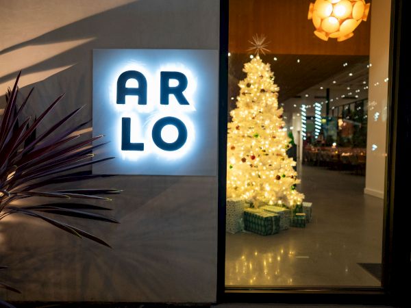 A nighttime storefront glows with an illuminated &ldquo;ARLO&rdquo; sign and a window display of lit Christmas trees and lights.