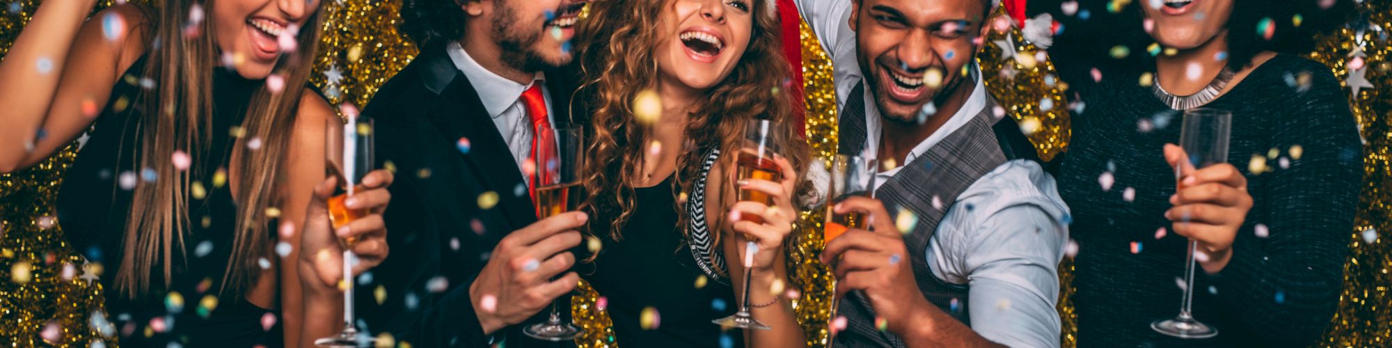A group of five people wearing Santa hats, holding drinks, and celebrating with confetti against a festive, glittery backdrop.