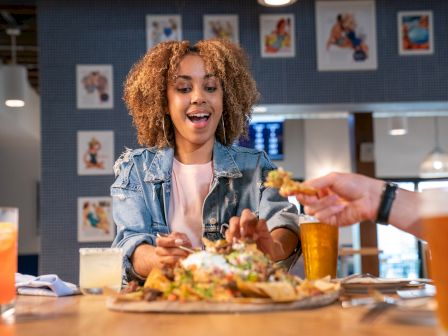 A person joyfully reaches for nachos at a restaurant, surrounded by drinks and colorful wall art in the background.