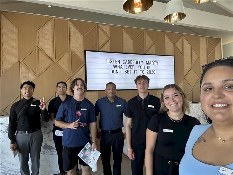 A group of six event staffers pose in front of a screen reading &ldquo;LISTEN CAREFULLY MARTY&hellip;&rdquo;, indoors.