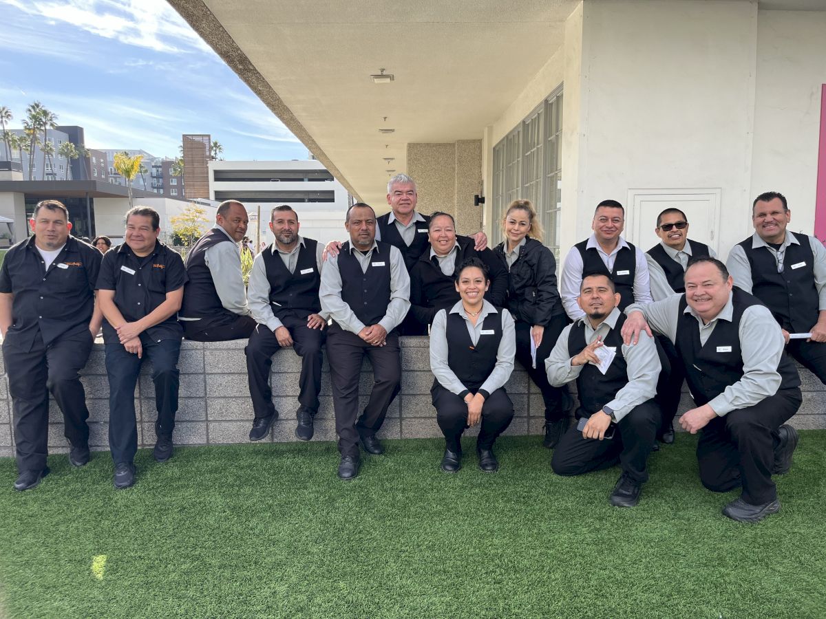 A group of coworkers in dark vests and white shirts pose together outdoors on the building steps, smiling for a photo.