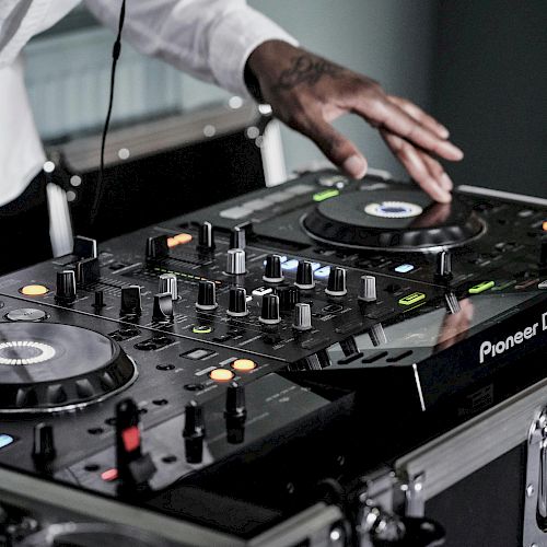A DJ is mixing music on a Pioneer DJ controller with various knobs and buttons being adjusted. The DJ's hand is on one of the jog wheels.