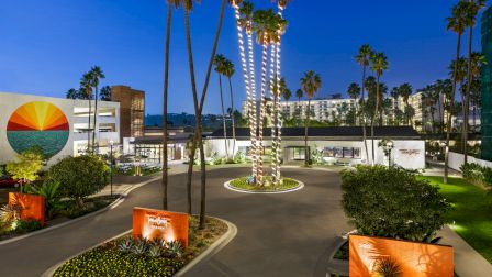 A nighttime resort courtyard with tall palm trees, glowing light strings, landscaped gardens, and modern hotel buildings.