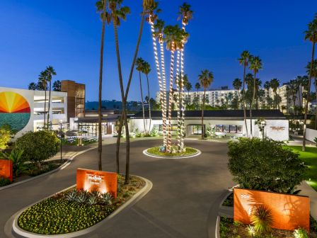 A nighttime resort courtyard with tall palm trees, glowing light strings, landscaped gardens, and modern hotel buildings.