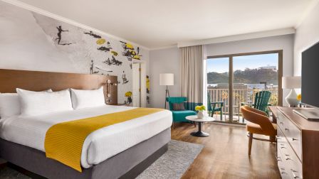 A modern hotel room with a large bed, teal chairs, desk, and balcony view, plus a street-art mural on the wall.