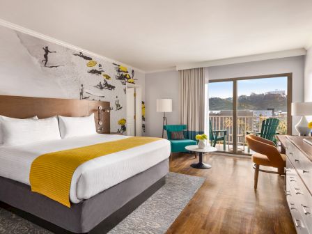 A modern hotel room with a large bed, teal chairs, desk, and balcony view, plus a street-art mural on the wall.