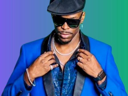 A person in a blue suit and hat, wearing sunglasses and jewelry, poses against a colorful gradient background.