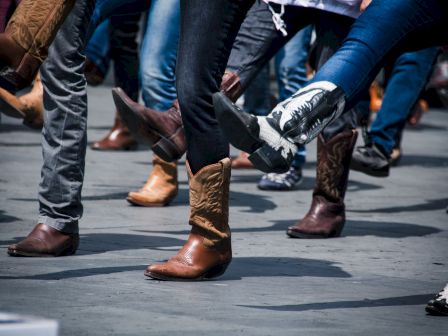 People are line dancing, wearing various styles of cowboy boots and jeans, captured mid-step on a gray surface.