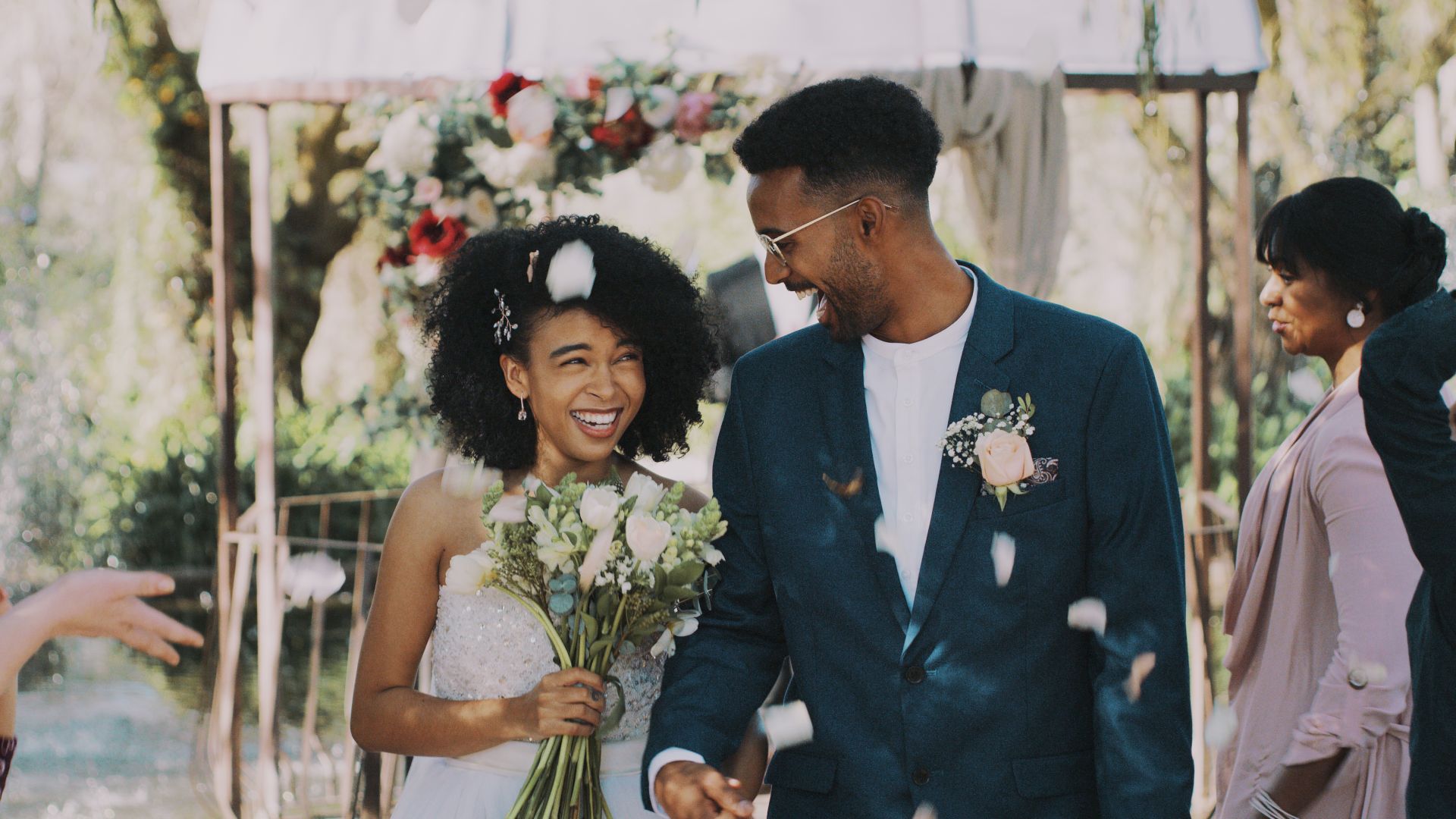 A couple smiles joyfully during an outdoor wedding ceremony, surrounded by guests and flower petals, under a decorated canopy.