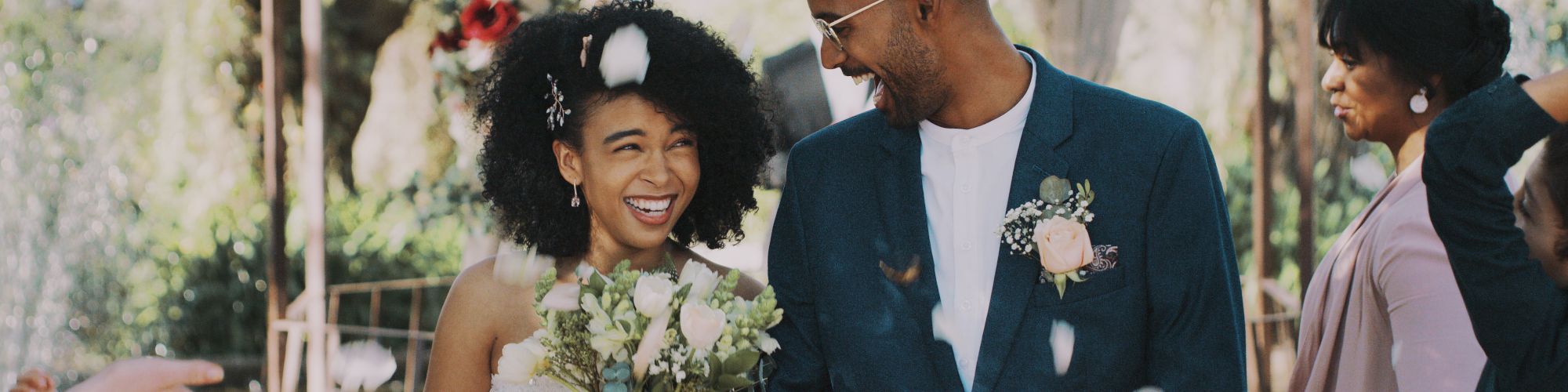 A couple smiles joyfully during an outdoor wedding ceremony, surrounded by guests and flower petals, under a decorated canopy.