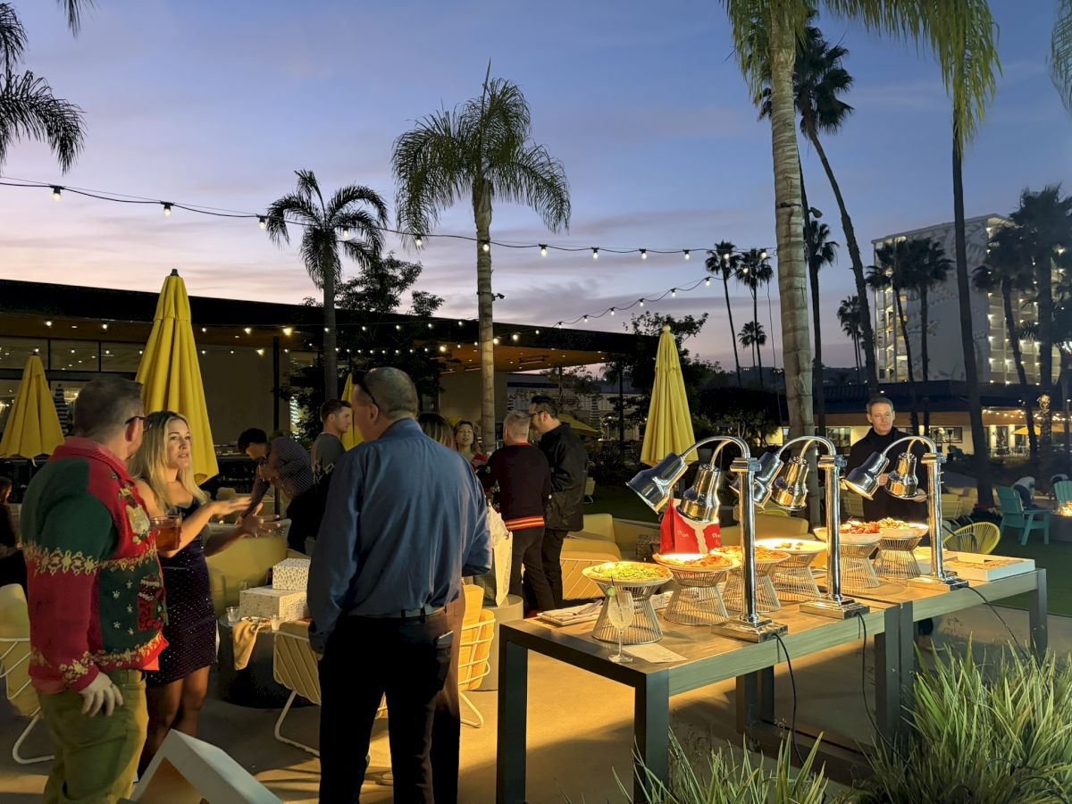 Outdoor happy-hour buffet with beer taps, string lights, palm trees, and guests chatting at dusk.