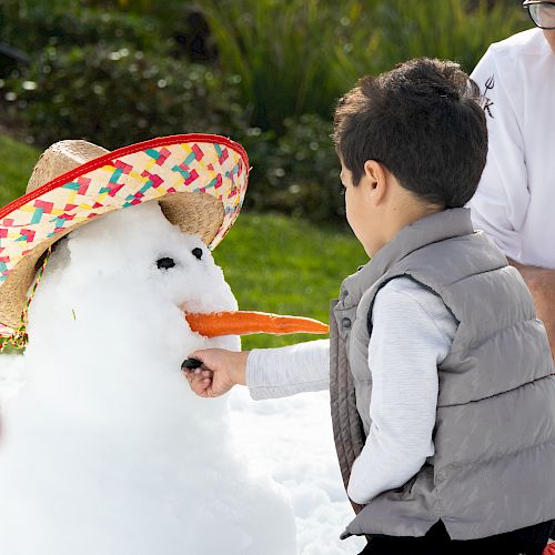 A child places a button on a snowman with a carrot nose and a sombrero hat, while a person in white appears partially in the background.