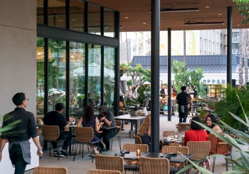 An outdoor dining area with people seated at tables, surrounded by plants, having meals while a waiter walks by.