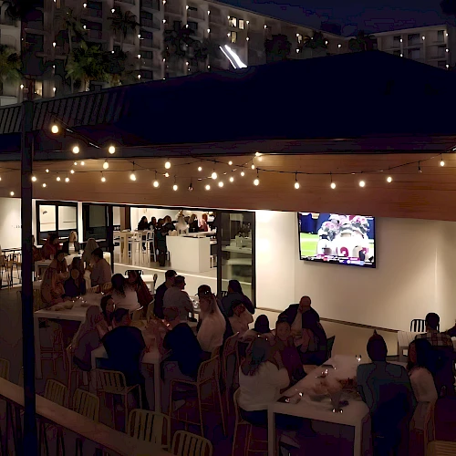 An outdoor dining area at night with string lights, tables of people, and a TV screen showing a sports game.