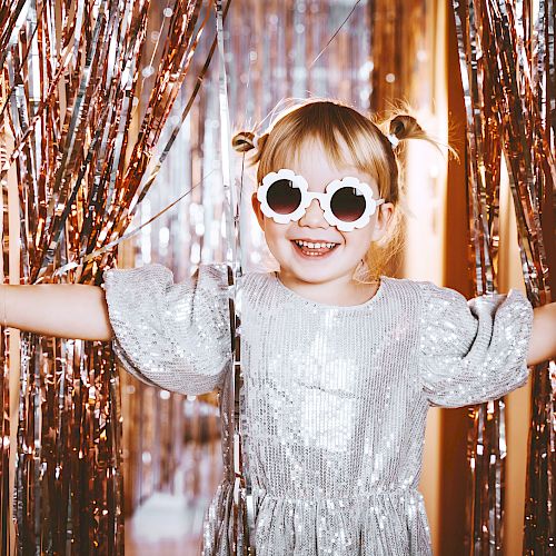 A child in a sparkly dress and sunglasses smiles, surrounded by metallic streamers, creating a festive and joyful scene.