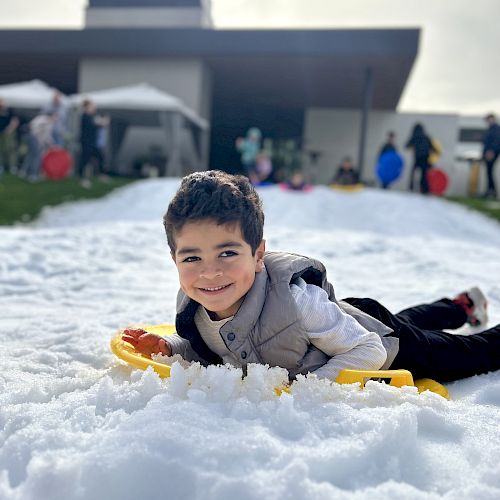 A child is lying on snow with a sled, smiling, in front of a modern building.