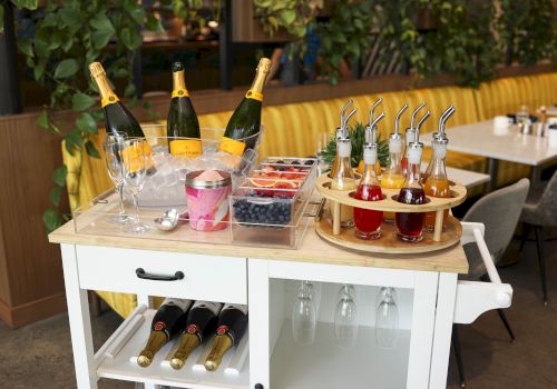 A white cart with champagne bottles, glasses, berries, and syrup bottles with straws arranged in a dining area.