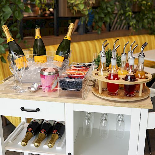 A white cart with champagne bottles, glasses, berries, and syrup bottles with straws arranged in a dining area.