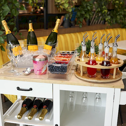 A white cart with champagne bottles, glasses, berries, and syrup bottles with straws arranged in a dining area.