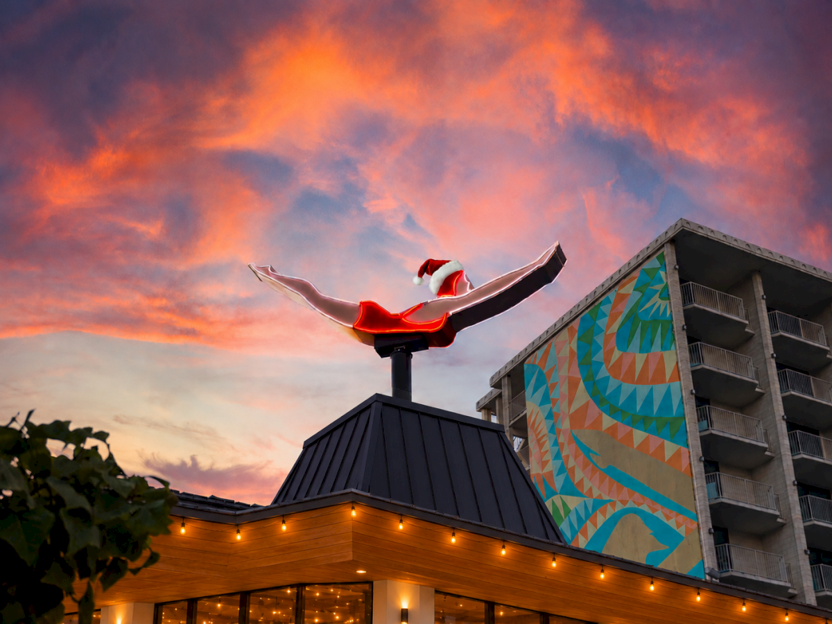 A colorful winged statue rises above a rooftop next to a vivid mural building under a glowing sunset sky.