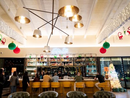 A festive bar interior with brass pendant lights, glowing string lights, holiday d&eacute;cor, cocktails, and people sitting at the counter, in a cheery gathering spot.