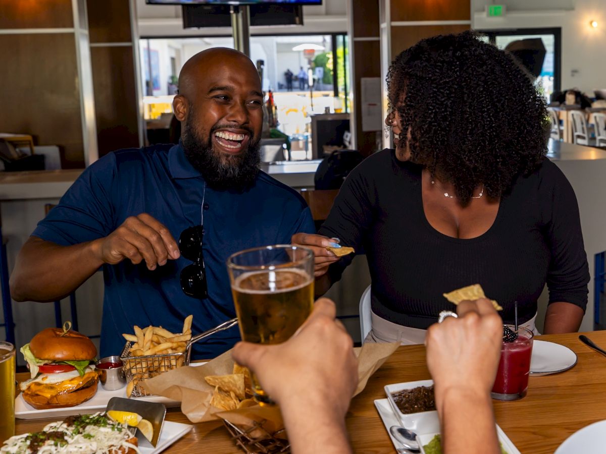 A smiling couple dine at a restaurant, enjoying burgers, fries, and drinks at a table.