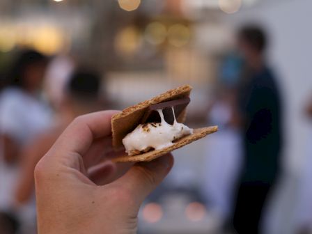 A hand holding a s’more with melted chocolate and marshmallow between graham crackers, with people blurred in the background.