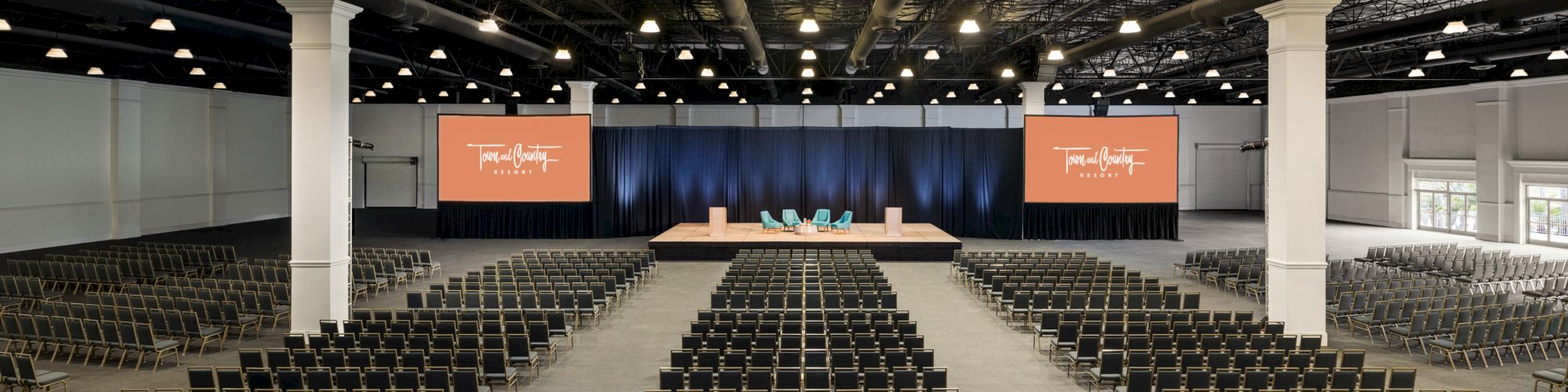 The image shows a large conference hall with rows of chairs facing a stage with screens and "This is the time" displayed.