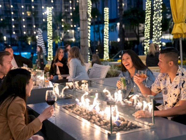 A group of people are enjoying drinks around a modern outdoor fireplace at night, with decorated trees and lights in the background.