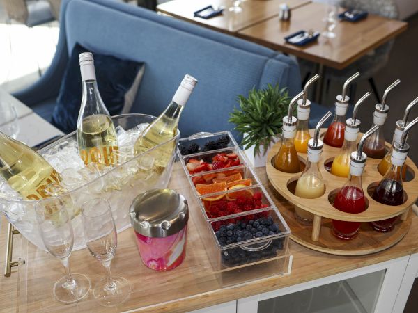 A cocktail drink station with bottles, glasses on ice, fruit bowls, and colorful juice dispensers on a wooden tray.