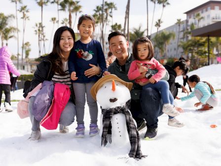 A family of four smiles in the snow, posing with a snowman on a playground while other kids play nearby.