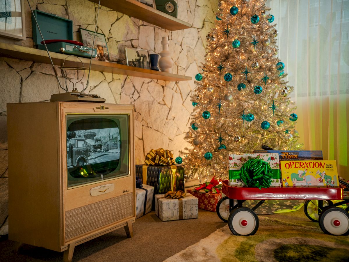 A decorated Christmas room with a lit tree, blue ornaments, wrapped gifts, a vintage TV, and a wooden toy-style truck in front.