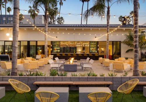 Outdoor lounge area with string lights, palm trees, modern seating, and a central fire pit in front of a bar.