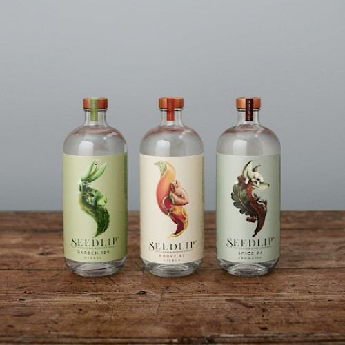 Three Seedlip bottles on a wooden surface, each with unique illustrated labels, set against a plain background.