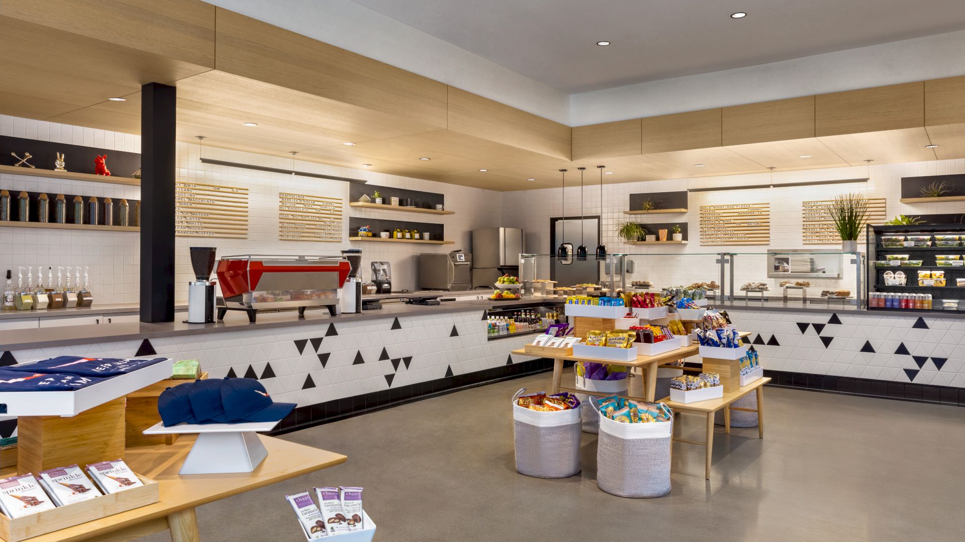 A modern cafe interior with a serving counter, shelves of snacks, coffee equipment, and neatly arranged tables displaying products.