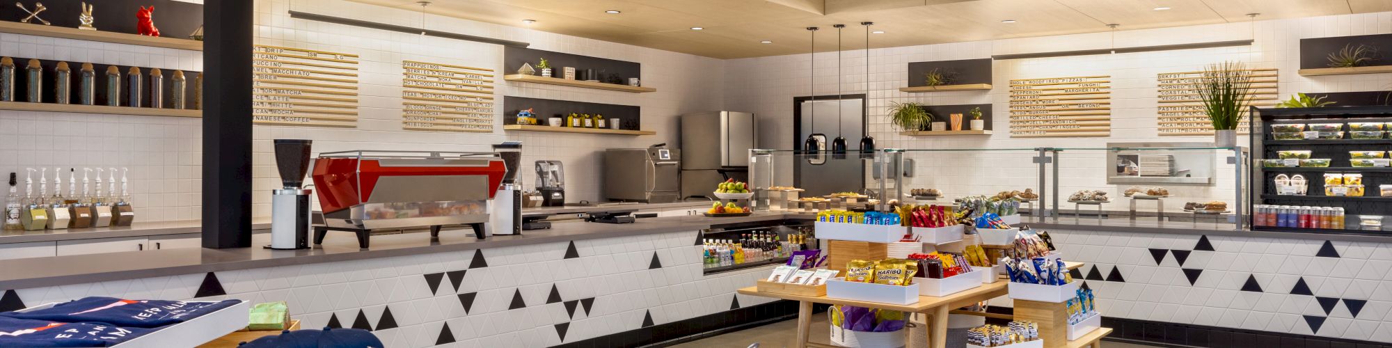 A modern cafe interior with a serving counter, shelves of snacks, coffee equipment, and neatly arranged tables displaying products.