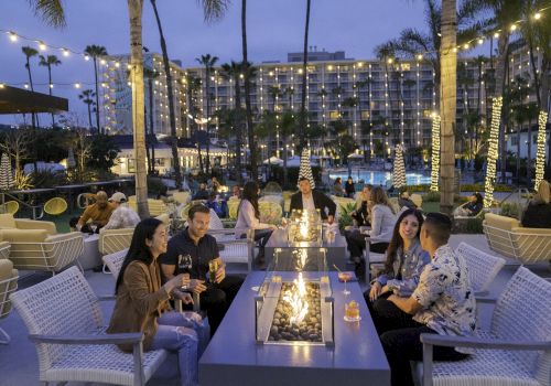 People are gathered around outdoor fire pits, enjoying drinks under string lights, with palm trees and a hotel in the background.