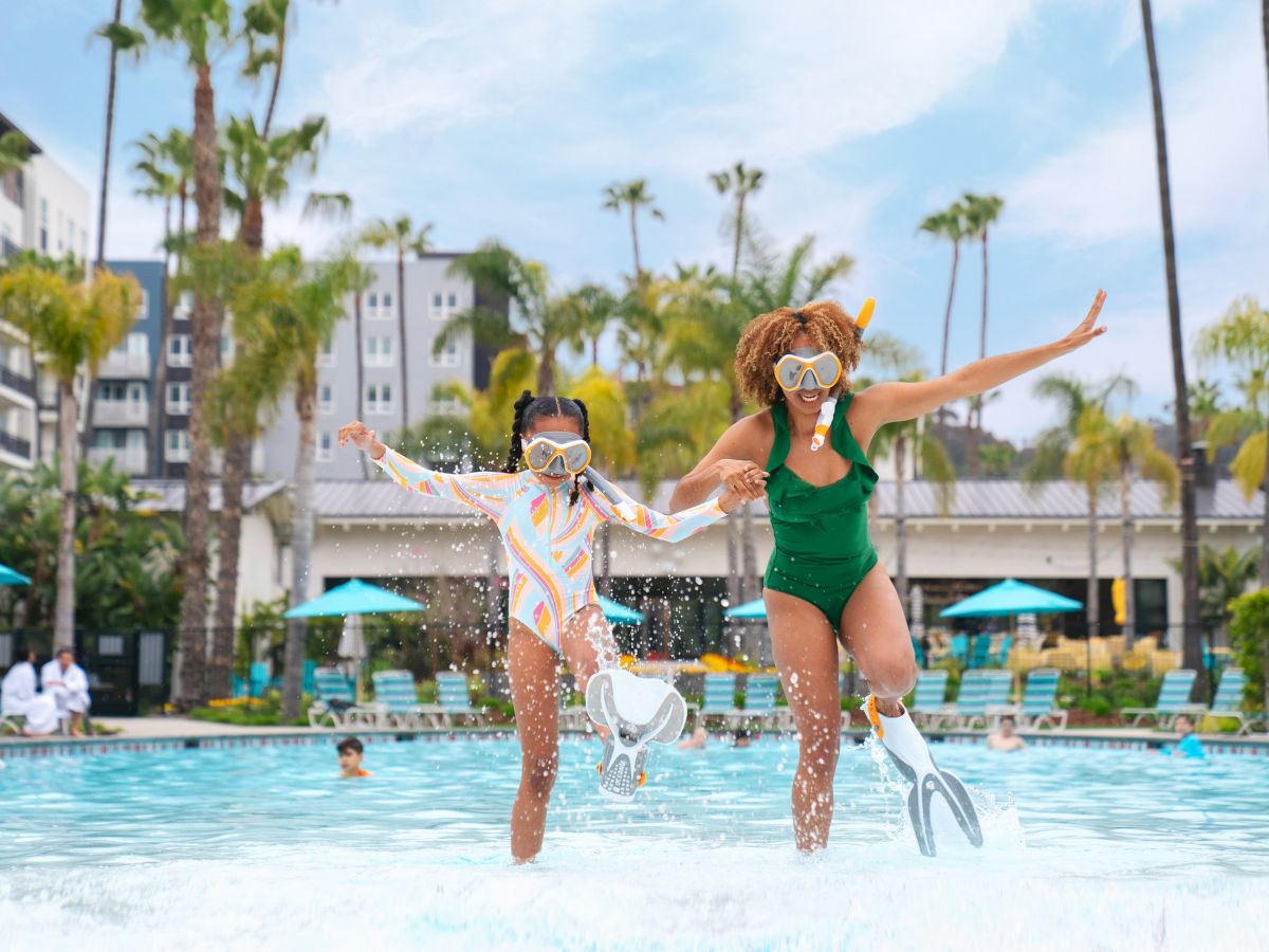Two people in swimsuits and goggles jump into a pool on a sunny day, with palm trees and buildings in the background.