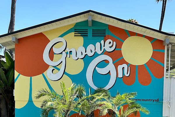 A colorful mural with "Groove On" text, featuring retro patterns and surrounded by tropical plants on a building wall.