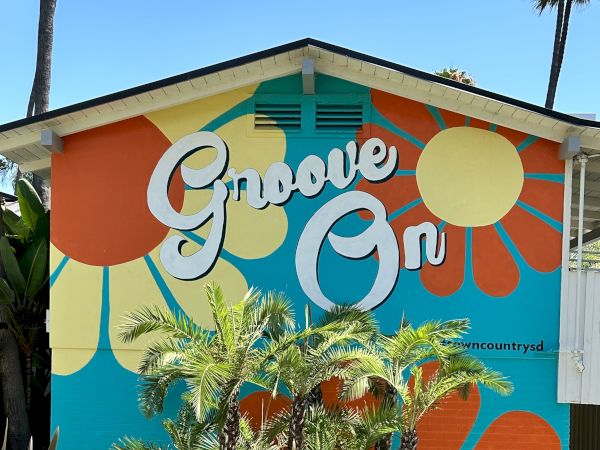 A colorful mural with "Groove On" text, featuring retro patterns and surrounded by tropical plants on a building wall.