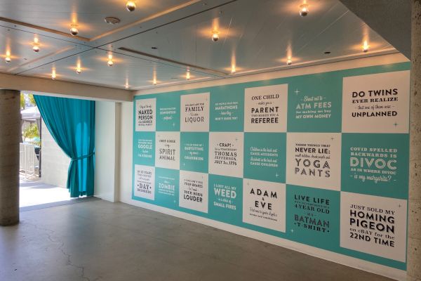 The image shows a wall with humorous quotes and sayings in a modern, well-lit room with blue curtains and ceiling lights.