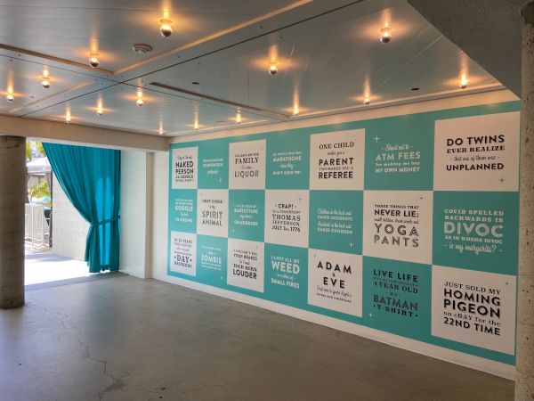 The image shows a wall with humorous quotes and sayings in a modern, well-lit room with blue curtains and ceiling lights.