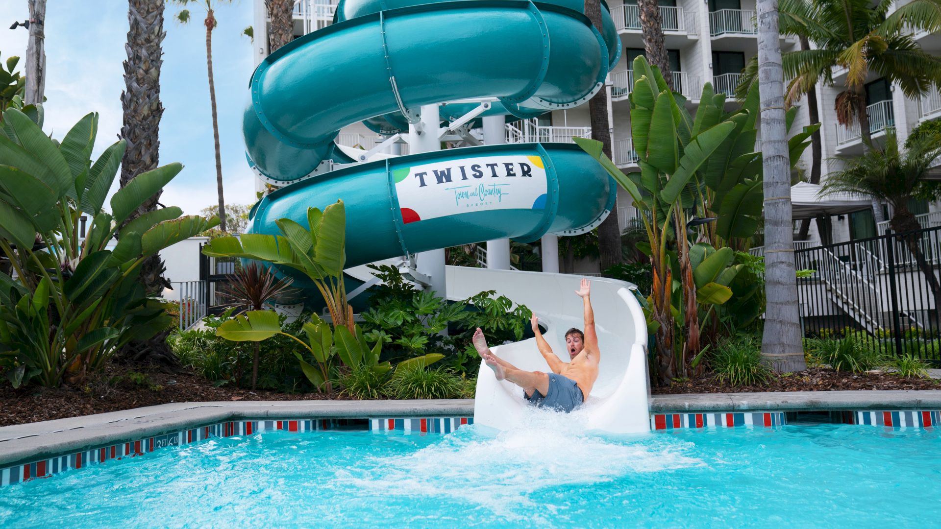 A person is sliding down a twisting water slide labeled "Twister" into a pool, with palm trees and a building in the background.