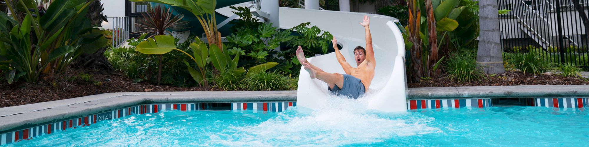 A person is sliding down a twisting water slide labeled "Twister" into a pool, with palm trees and a building in the background.